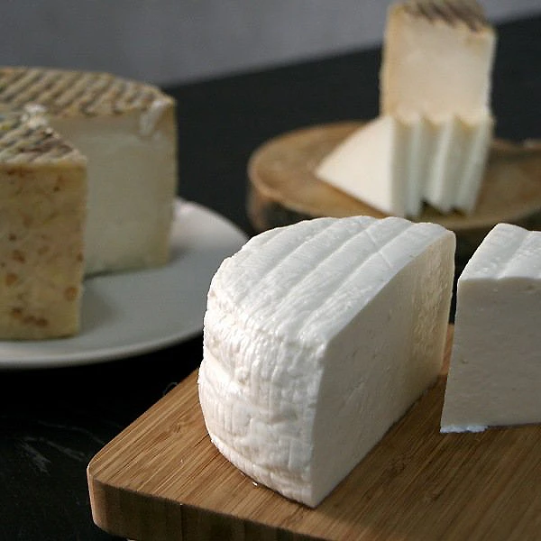 Queso de pasta blanda blanca sobre tabla de madera, con otros quesos detr&aacute;s.