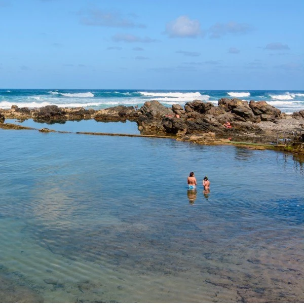 Dos personas nadan en aguas cristalinas junto a rocas y el mar.