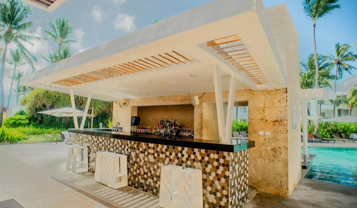 Bar al aire libre con techo blanco, palmeras y piscina al fondo, resort tropical.