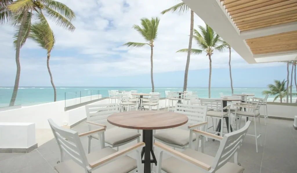 Terraza frente al mar con palmeras y mesas blancas bajo techo de madera.