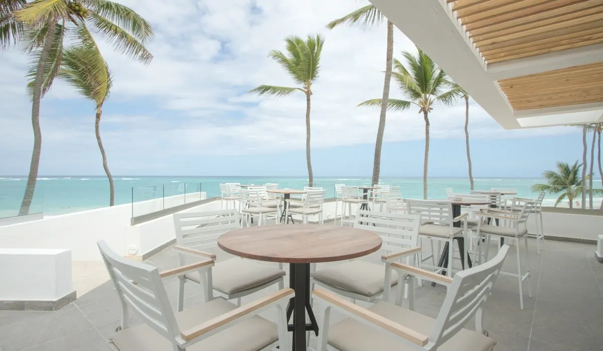 Terraza frente al mar con palmeras y mesas blancas bajo techo de madera.