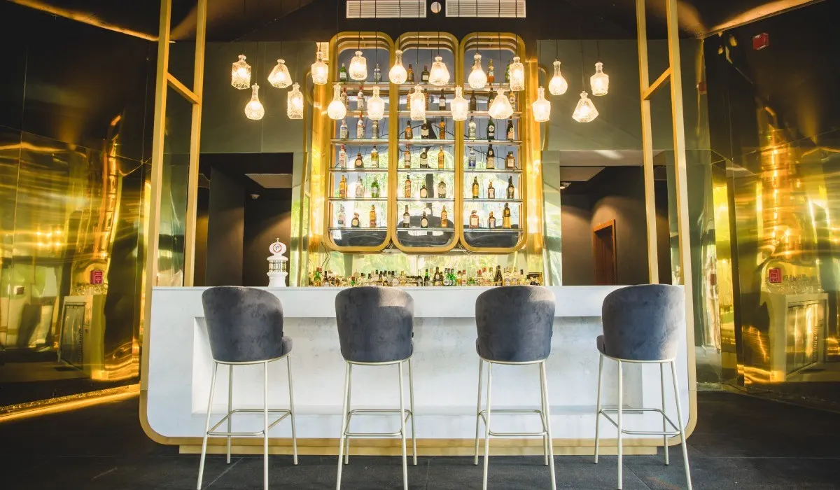 Bar iluminado con bebidas en exhibidor dorado y cuatro taburetes azules