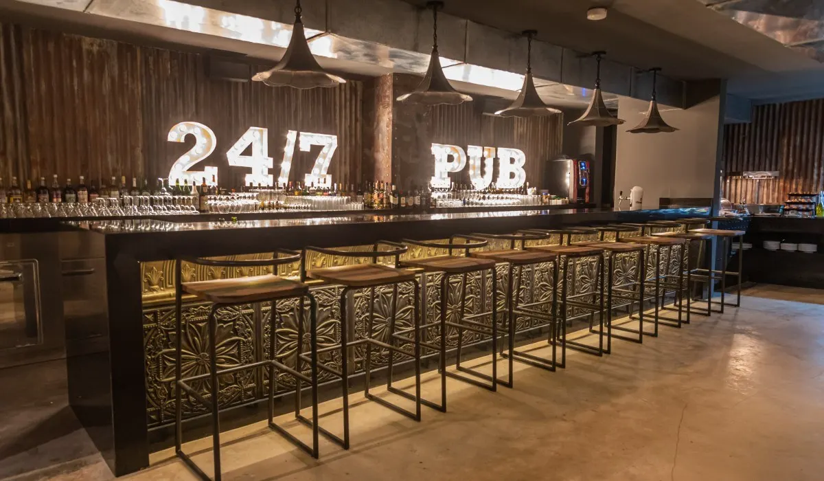 Bar con barra dorada, letreros &ldquo;24/7&rdquo; y &ldquo;PUB&rdquo;, vasos y taburetes.
