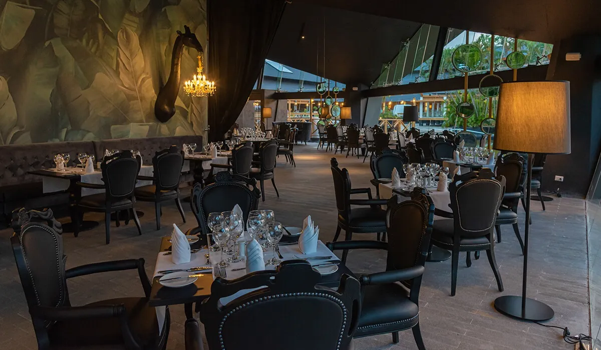 Comedor de restaurante con mesas servidas, sillas negras y l&aacute;mparas colgantes.