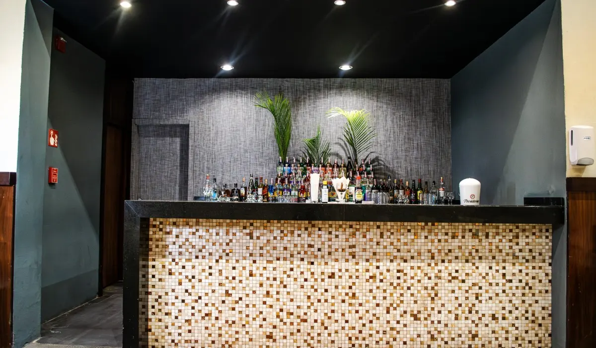 Bar con botellas y mosaico beige, plantas y luces en el techo.
