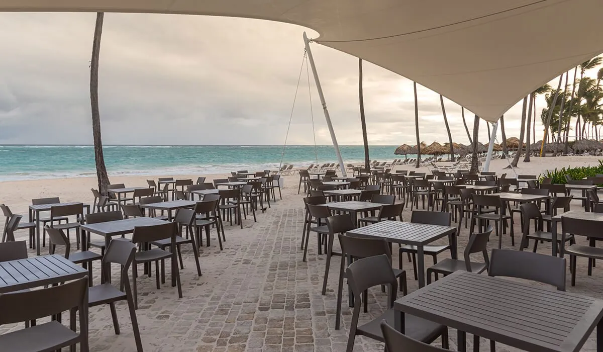 Terraza junto a la playa con mesas y sillas bajo una carpa blanca.