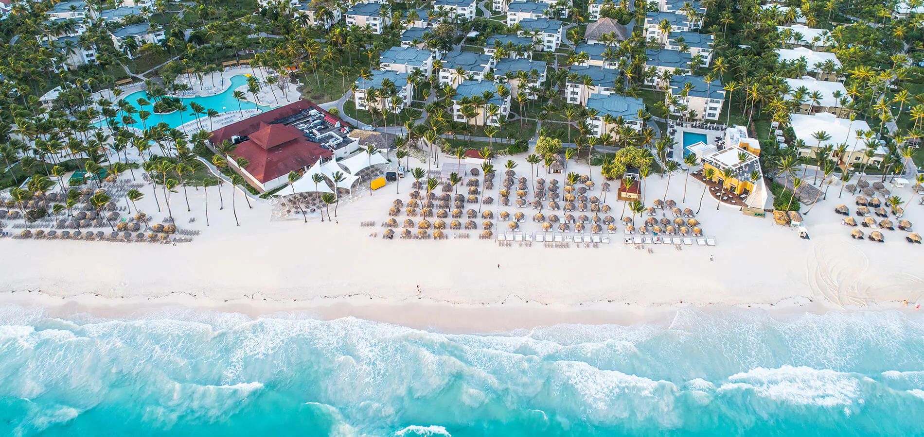 Vista a&eacute;rea de un resort con playa, palmeras, albercas y filas de mesas frente al mar