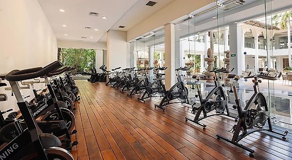 Gimnasio con bicicletas y m&aacute;quinas de ejercicio alineadas, luz natural y ventanas.