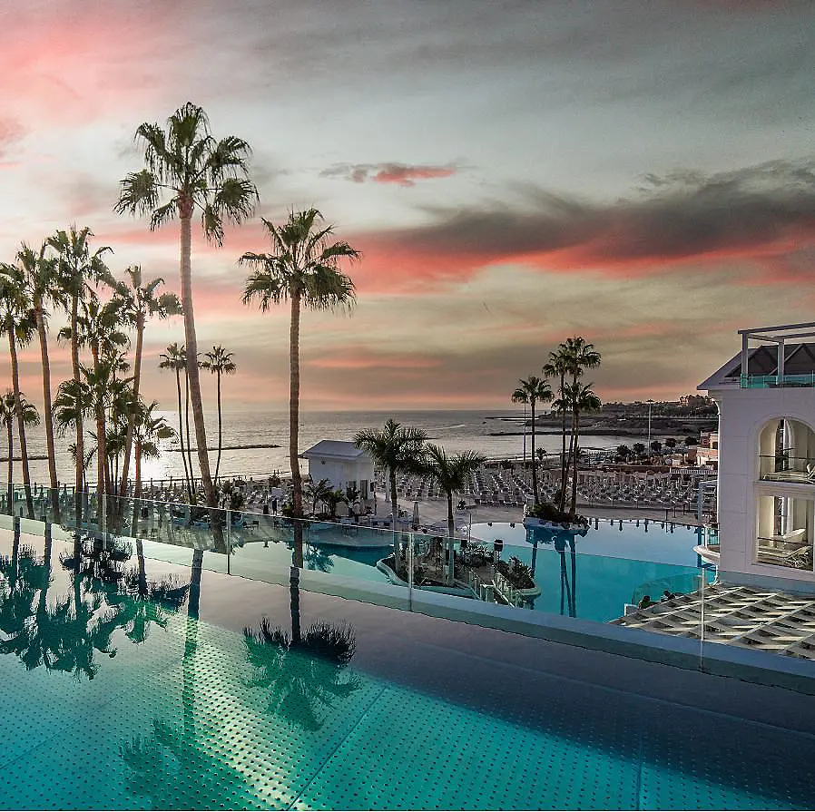 Piscina junto al mar al atardecer, palmeras y cielo con nubes rosadas