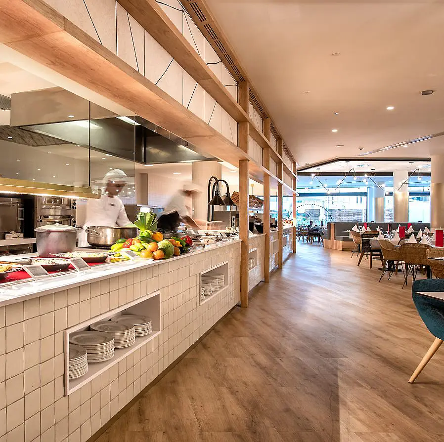 Cocina de hotel con estaci&oacute;n de comida y mesas al fondo, comedor.