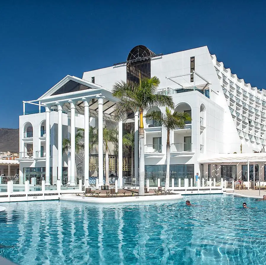 Hotel blanco moderno con piscina y palmeras bajo cielo azul