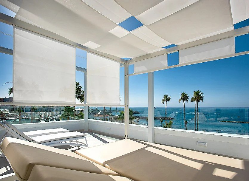 Terraza blanca con sof&aacute;s y vista al mar, palmeras y cielo azul