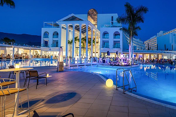 Piscina junto a edificio blanco con columnas y palmeras, de noche.