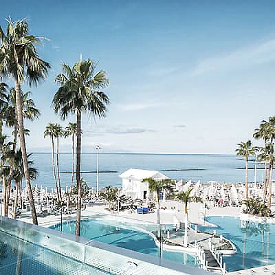 Piscina y palmeras junto al mar, con caba&ntilde;as blancas frente a la costa