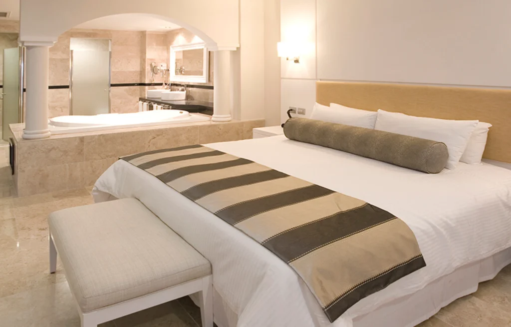 Suite con cama blanca, banco y ba&ntilde;o de m&aacute;rmol al fondo, estilo hotel.