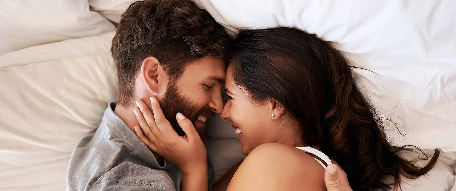 Pareja en la cama se besa y sonr&iacute;e, abrazados bajo s&aacute;banas blancas.