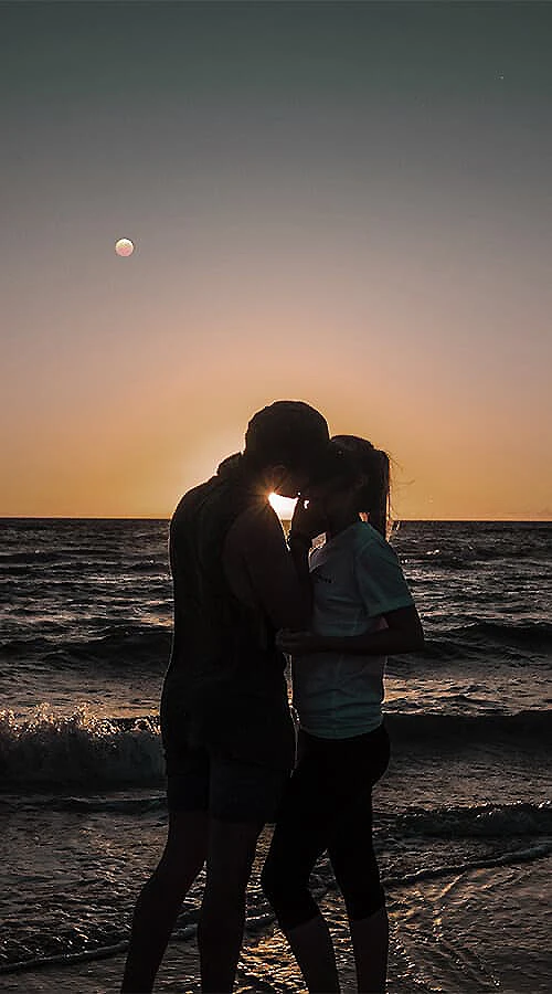 Pareja abrazada en la playa al atardecer, con luna en el cielo