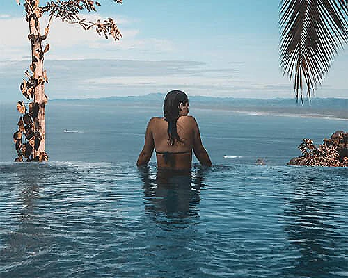 Mujer de espaldas en la piscina mirando el mar y el paisaje al atardecer
