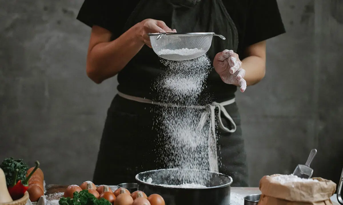 Cocinero tamiza harina sobre un bol con ingredientes en la mesa.