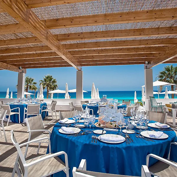 Mesa con vajilla y manteles azules en terraza frente al mar bajo p&eacute;rgola de madera