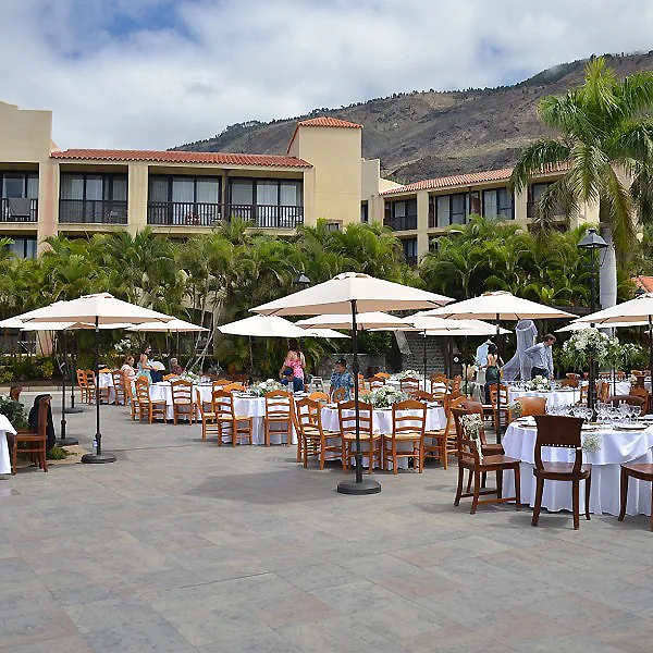 Patio de hotel con mesas, sillas y sombrillas blancas junto a palmeras y monta&ntilde;a