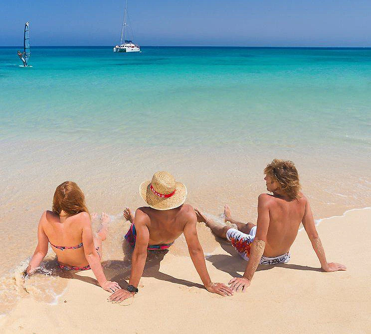 Tres personas en la playa, mirando al mar con barcos al fondo.