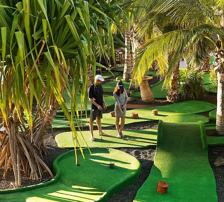 Dos personas juegan en un campo de minigolf entre palmeras y c&eacute;sped verde.