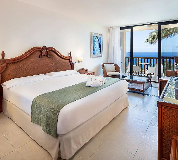 Dormitorio con cama y balc&oacute;n con vista al mar, silla y muebles de madera