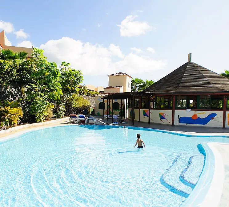 Piscina azul en un complejo con palmeras y edificios al fondo, bajo el sol.