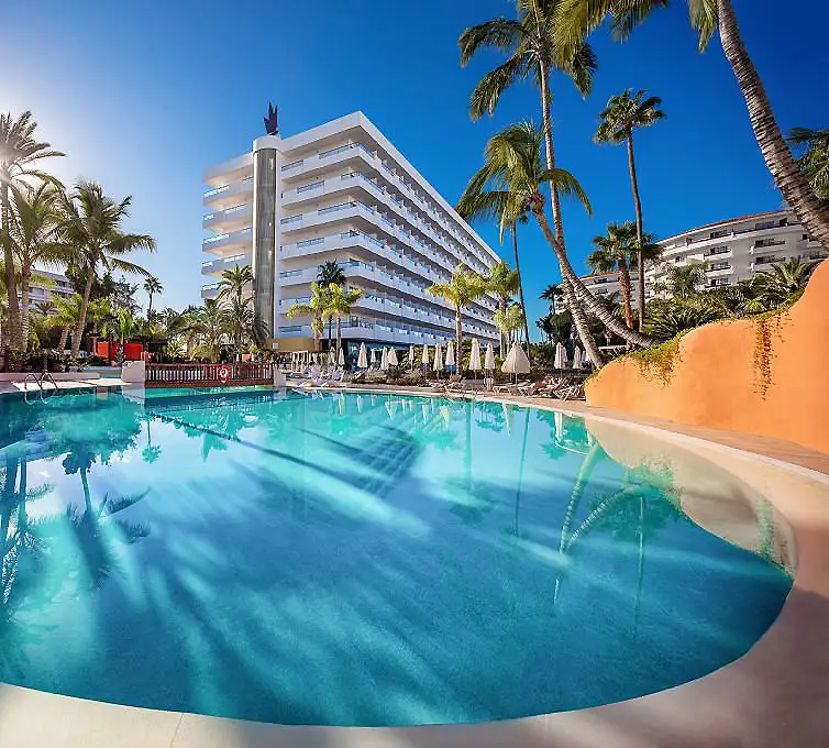 Piscina con palmeras y hoteles frente al mar, cielo azul tropical.