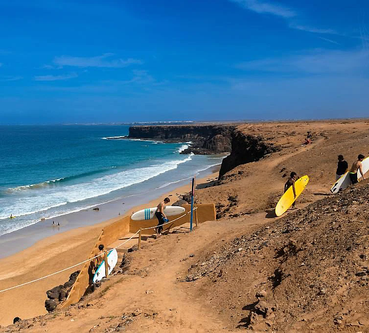 Playa con surfistas y tabla amarilla junto a acantilado y mar azul.