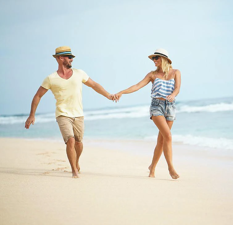 Pareja paseando de la mano en la playa, con sombreros y ropa casual.