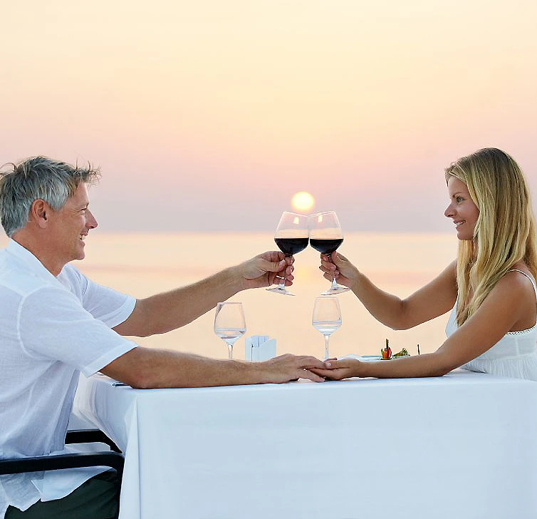 Pareja brinda con copas de vino al atardecer, en una mesa junto al mar.