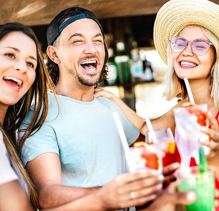 Tres amigos brindan con bebidas en una feria o bar, sonriendo y riendo.