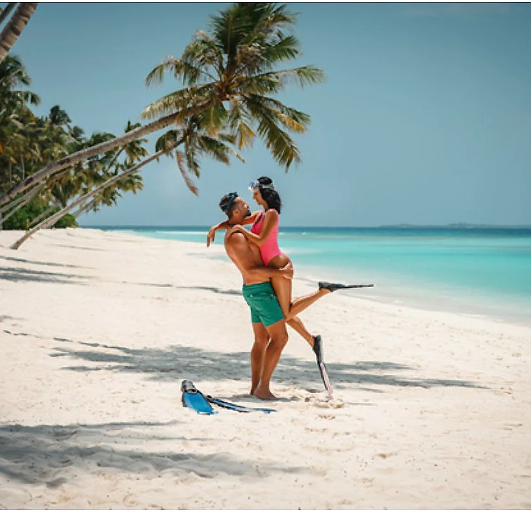 Pareja en la playa tropical abrazada, con palmeras y olas al fondo.