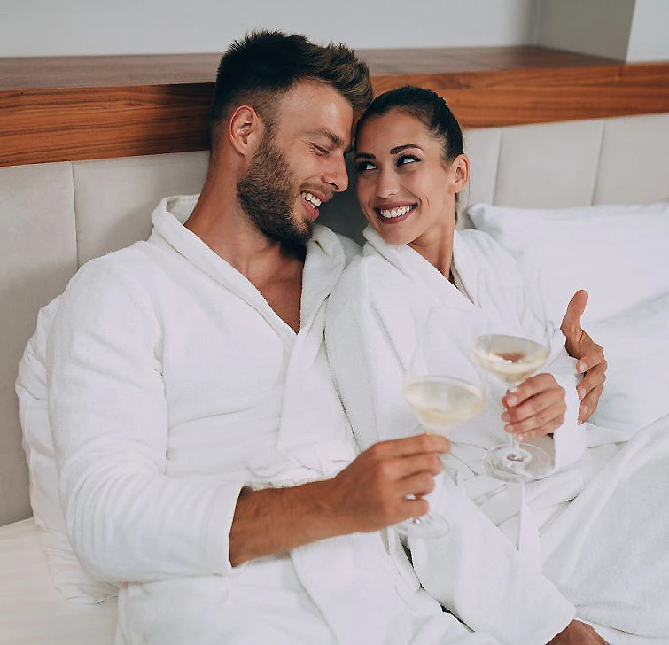 Pareja en batas blancas en cama brindando con copas de vino.