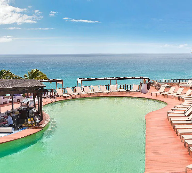 Piscina al borde del mar con tumbonas y vista al oc&eacute;ano, cielo despejado.
