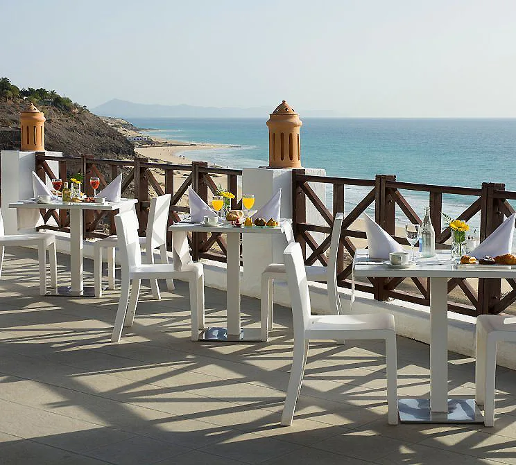 Mesa con sillas y desayuno en terraza frente al mar, con costa al fondo.