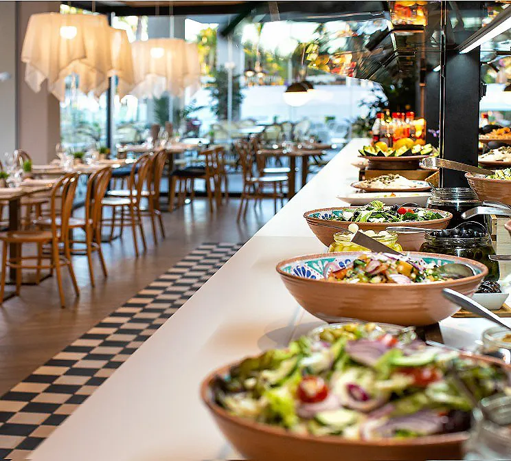 Buffet de ensaladas y platos en un restaurante con mesas y sillas al fondo.