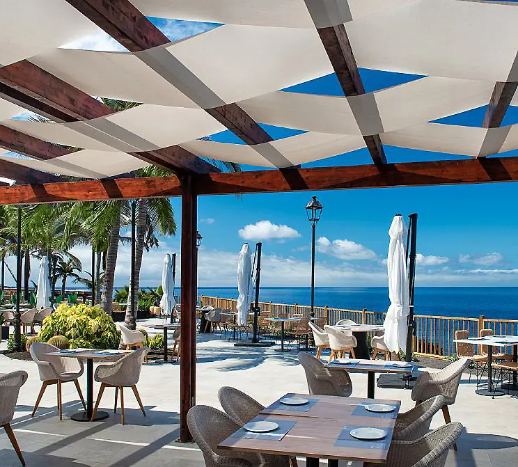 Terraza frente al mar con mesas, sillas y toldos blancos bajo palmeras.