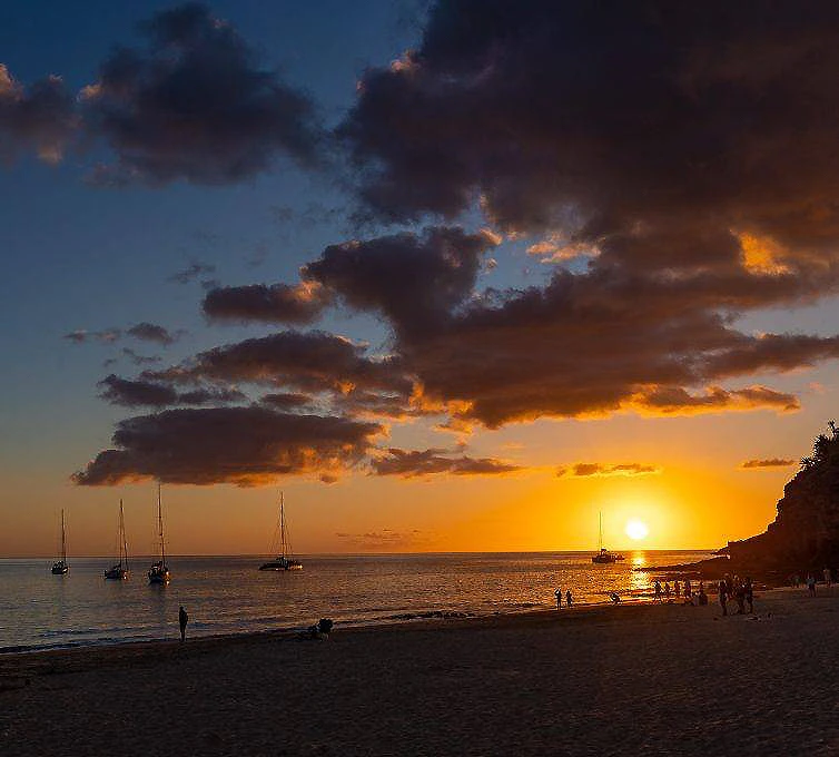 Puesta de sol en la playa con barcos al fondo y nubes dram&aacute;ticas.