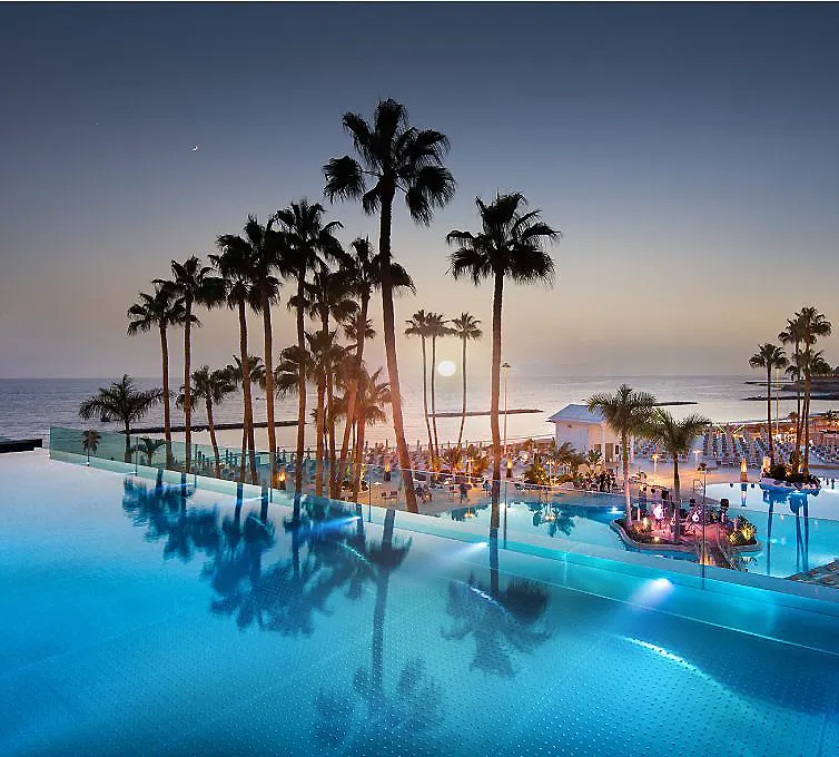 Piscina de hotel frente al mar al atardecer, con palmeras y luz azul