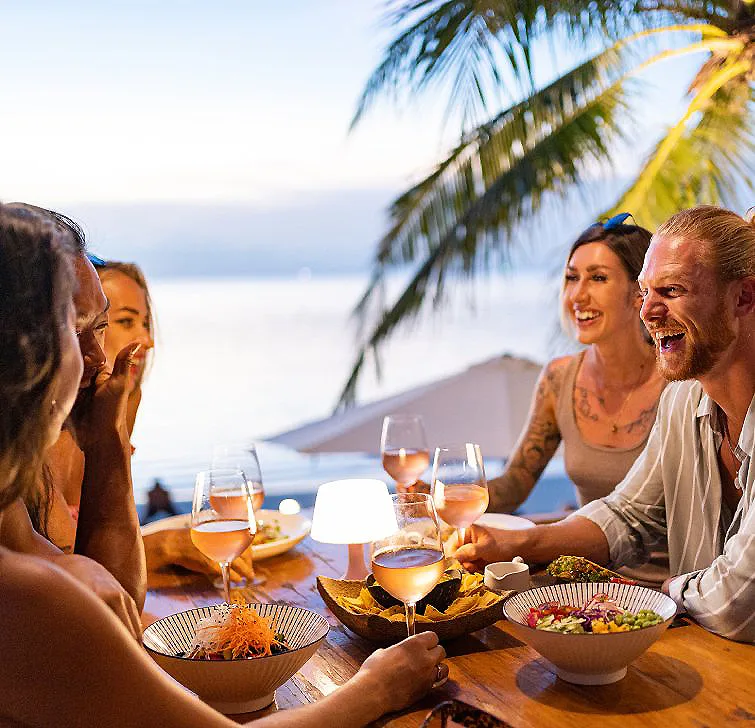 Amigos brindando y riendo en la mesa de una cena en la playa al atardecer.