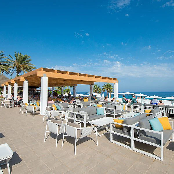 Terraza frente al mar con tumbonas y mesas bajo p&eacute;rgola, cielo despejado.