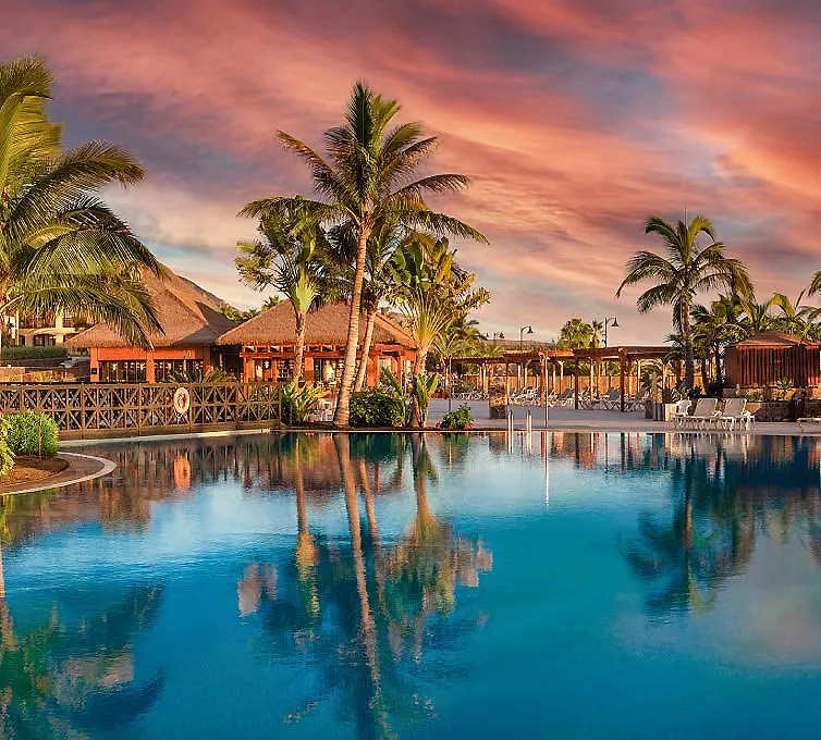 Piscina tropical con palmeras y caba&ntilde;as al atardecer, cielo rosado.