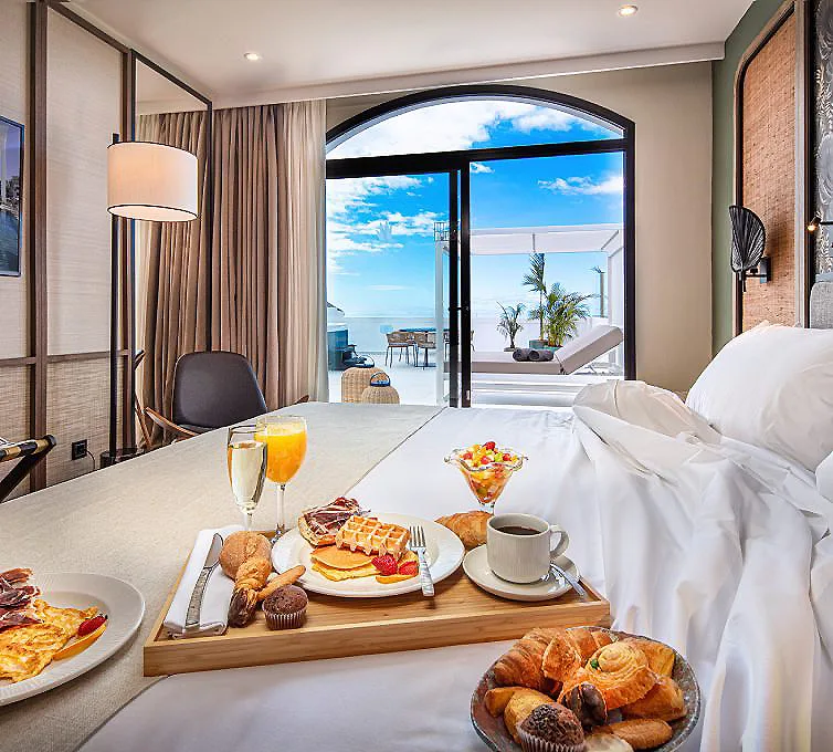 Desayuno en mesa con croissants, fruta y caf&eacute; frente a vista al mar.