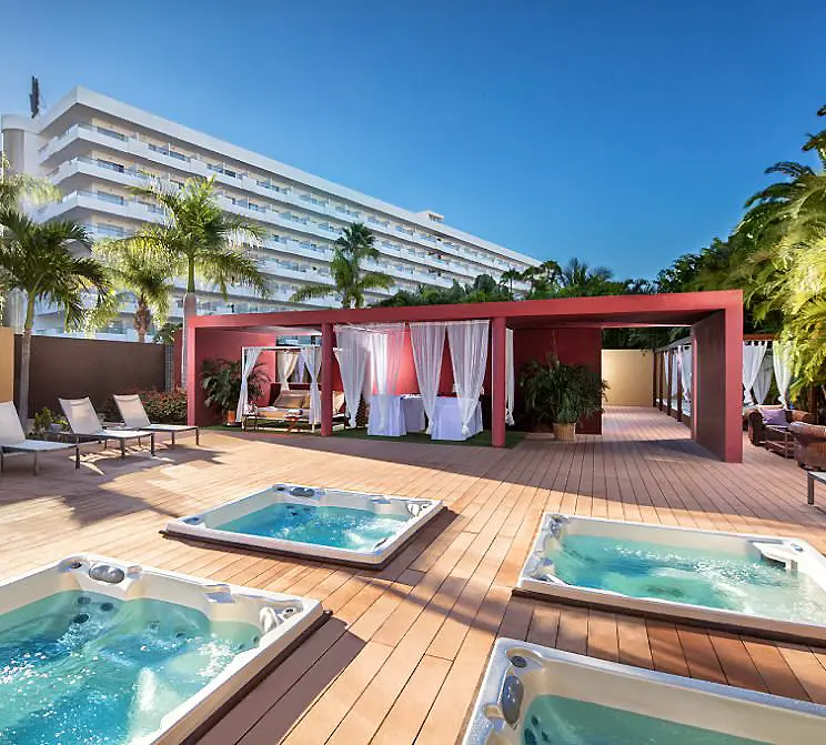 Piscina y jacuzzi al aire libre junto a hotel con palmeras y cielo azul
