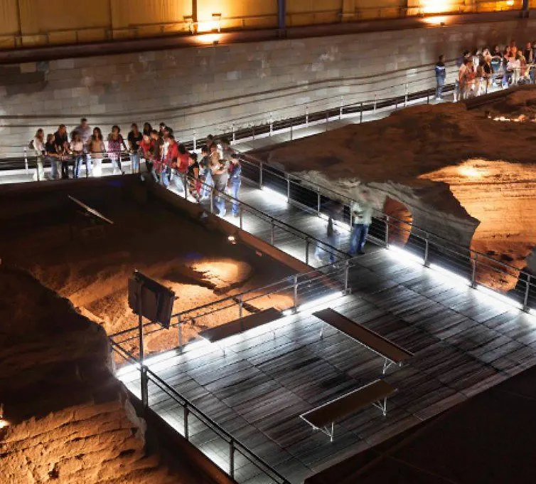 Turistas caminan en un pasillo elevado junto a una excavaci&oacute;n arqueol&oacute;gica.