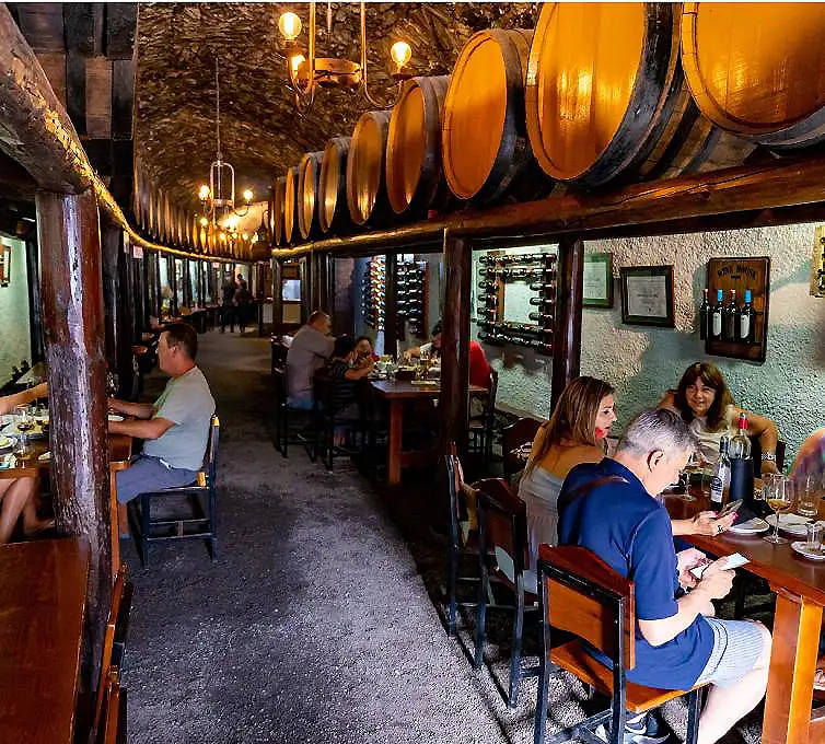 Interior de bodega con barricas de vino y clientes cenando en mesas.