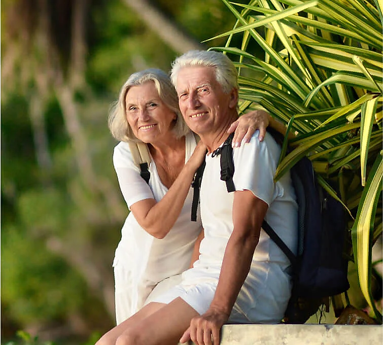 Pareja mayor sonriendo junto a una palmera, al aire libre.